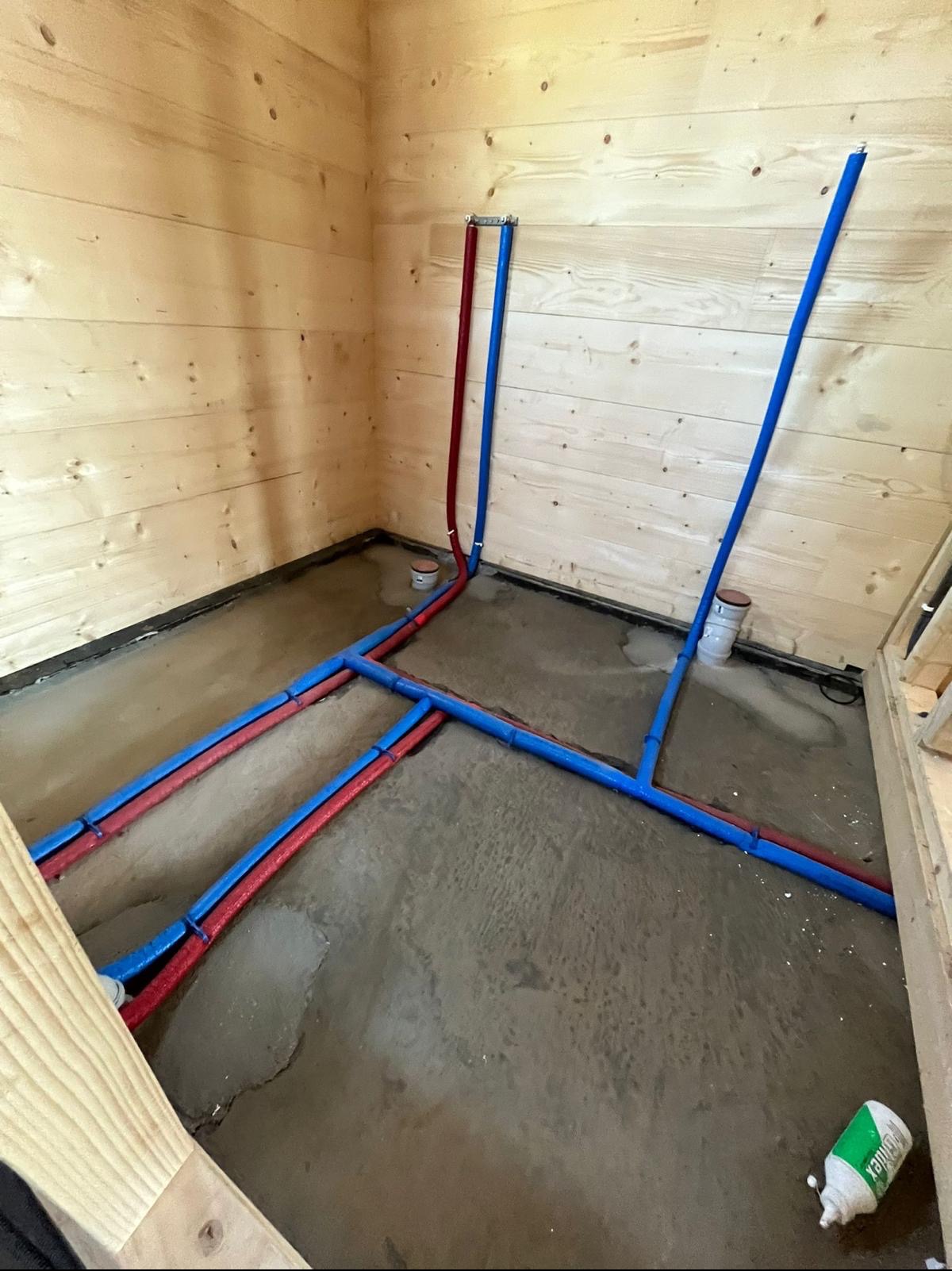 Wnętrze pomieszczenia z drewnianymi ścianami, w którym ułożono rury instalacji wodnej w kolorach czerwonym i niebieskim, częściowo zatopione w szarej wylewce betonowej. Widoczny fragment drewnianej...