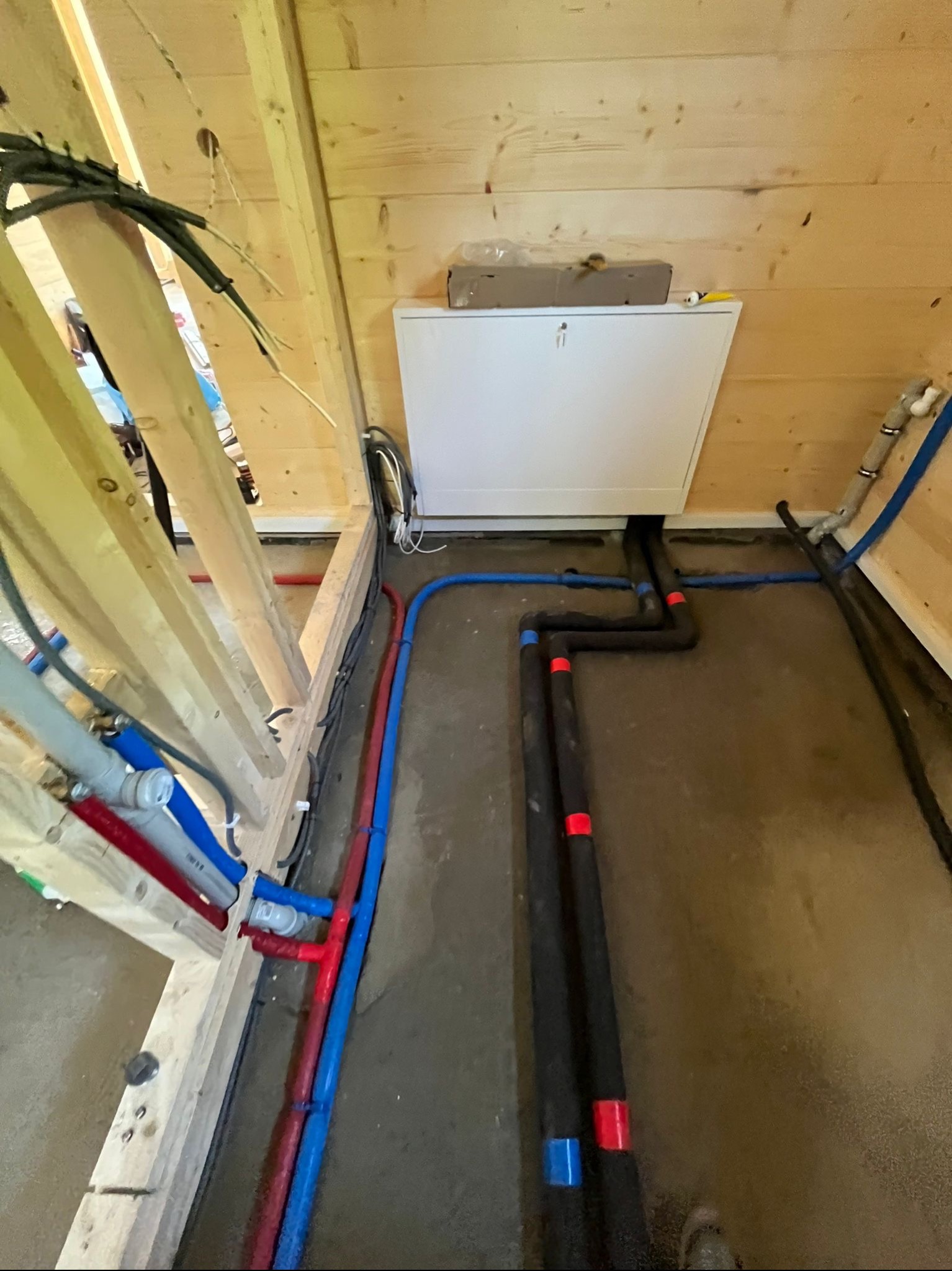 Wnętrze drewnianego domu w trakcie budowy z widocznymi instalacjami: rurami wodnymi w kolorach niebieskim i czerwonym, czarnymi rurami izolowanymi termicznie oraz białą skrzynką rewizyjną na ścianie.