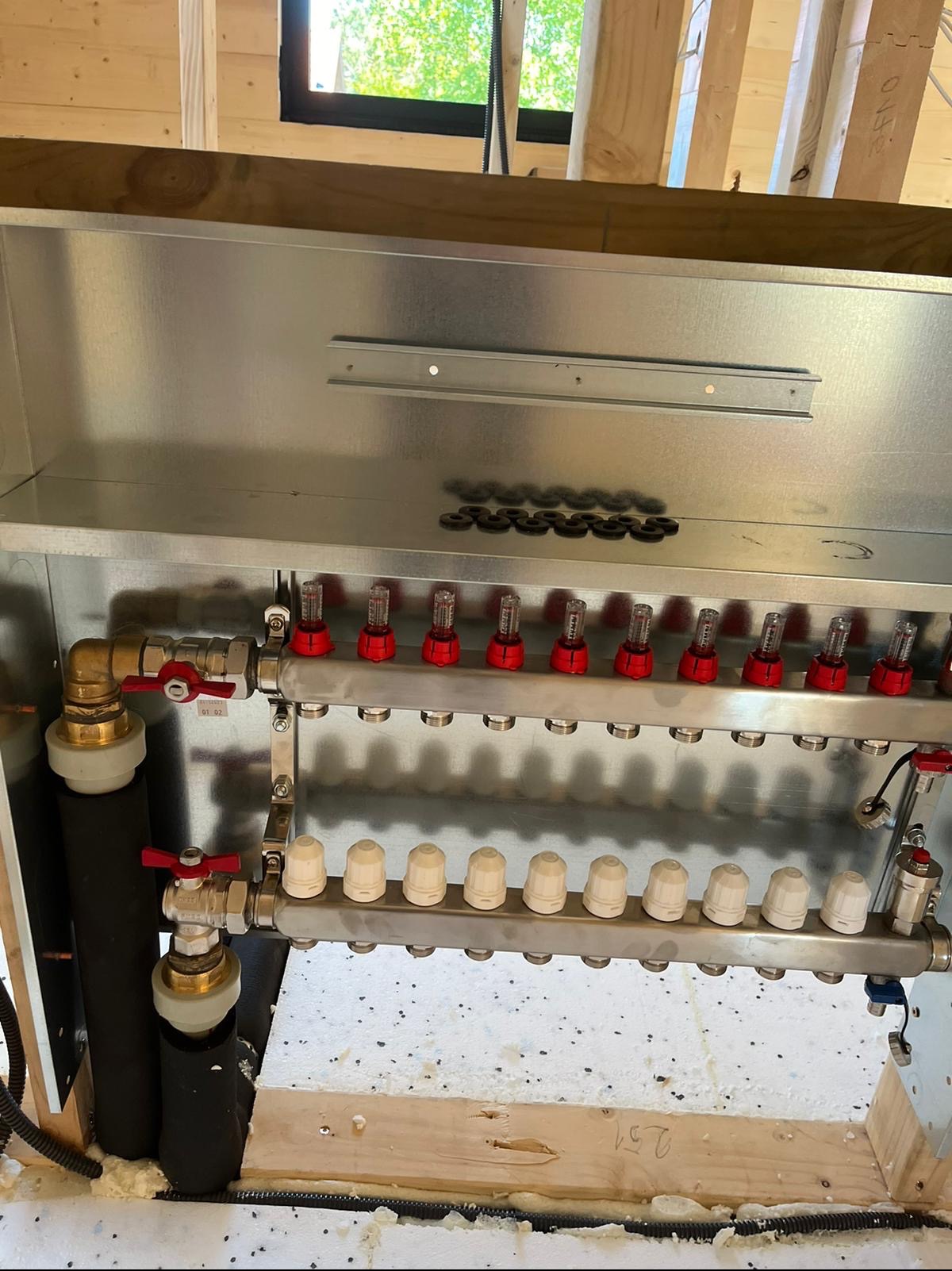 Rozdzielacz ogrzewania podłogowego z odpowietrznikami, zamontowany w skrzynce, widoczne przyłącza i izolacja, surowe drewniane elementy konstrukcyjne w tle.