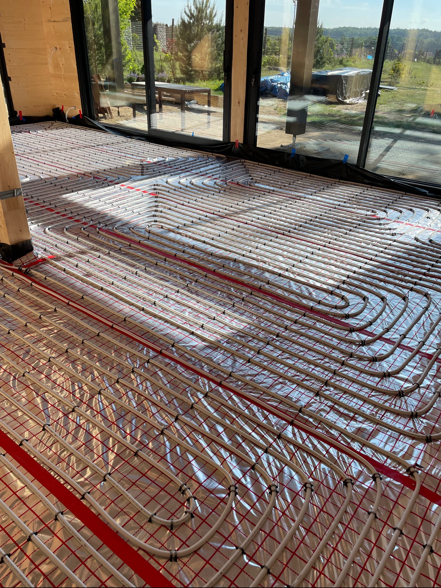 Białe rurki ogrzewania podłogowego ułożone na folii izolacyjnej z czerwoną siatką w nowym domu z dużymi oknami.