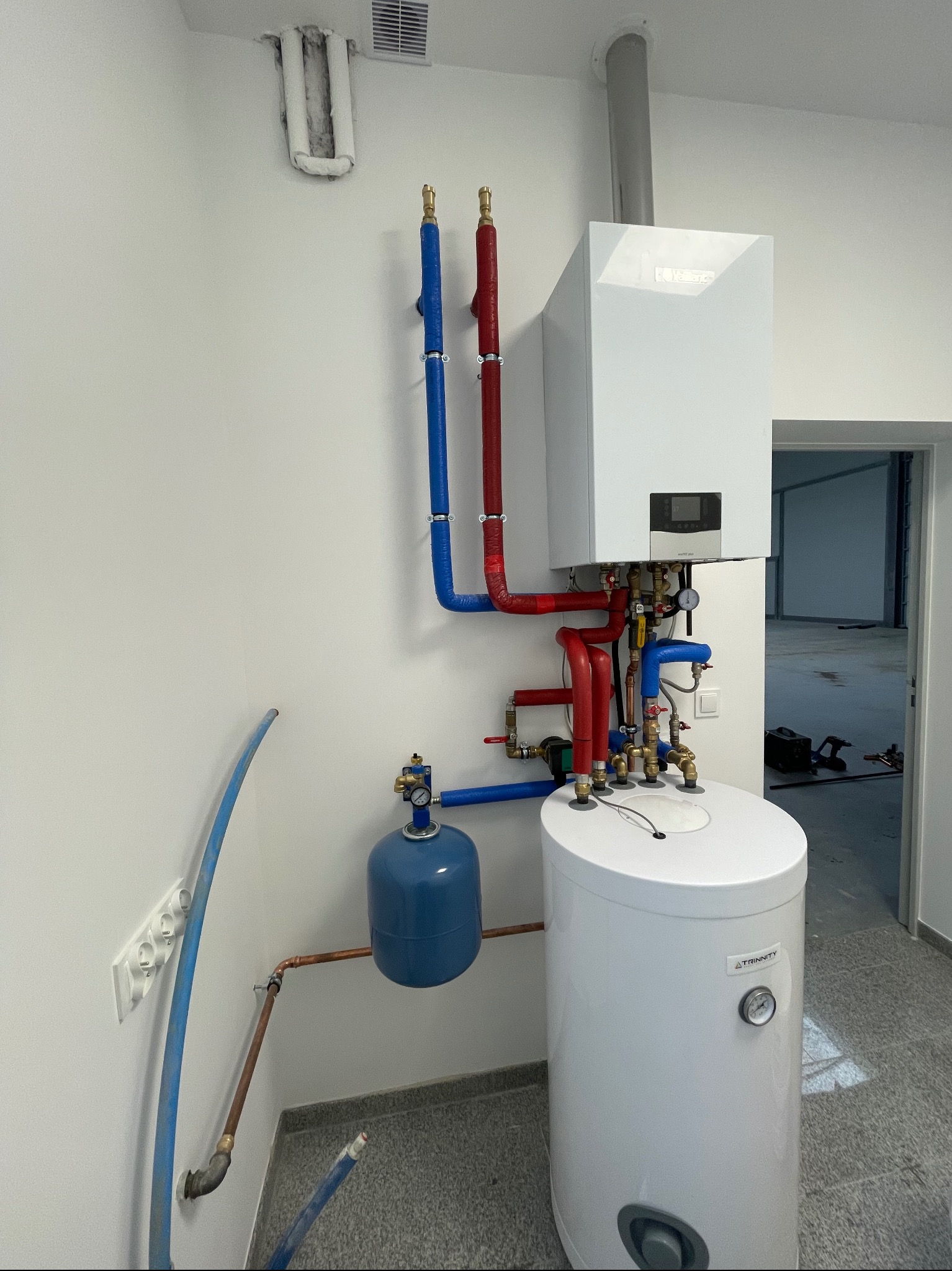 Biały piec gazowy Vaillant z podłączonym zasobnikiem wody Trinity, rurami w izolacji czerwonej i niebieskiej, oraz niebieskim naczyniem przeponowym, zamontowane na białej ścianie w nowym budynku...