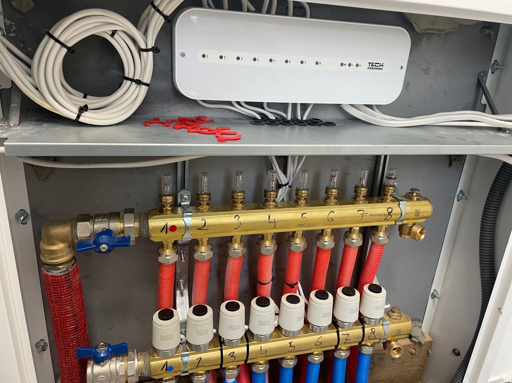 Rozdzielacz ogrzewania podłogowego z zainstalowanymi zaworami termostatycznymi, widoczne rury doprowadzające ciepłą wodę w kolorze czerwonym i odprowadzające w kolorze niebieskim, listwa sterująca...