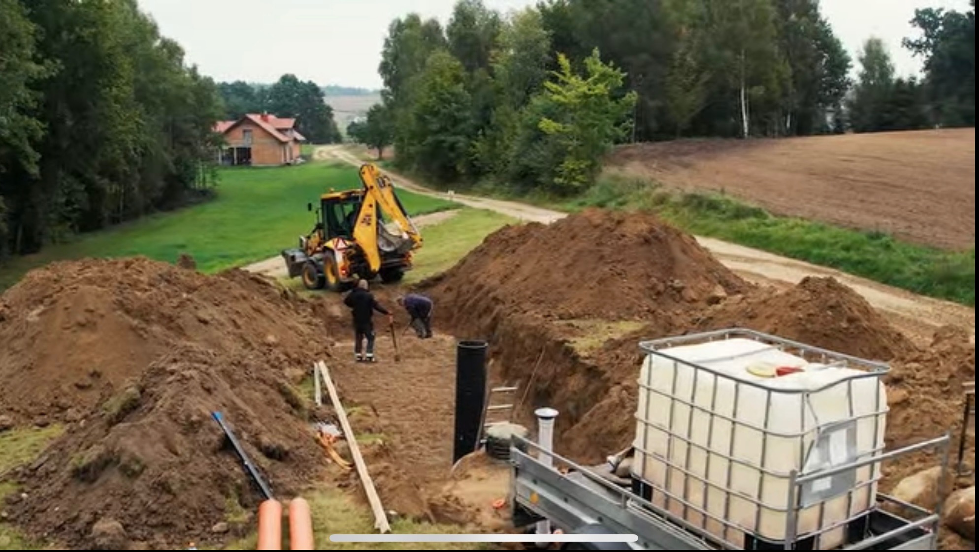 Wykonawca kopie rów koparką, przygotowując teren pod montaż zbiornika na ścieki na działce z widokiem na dom.