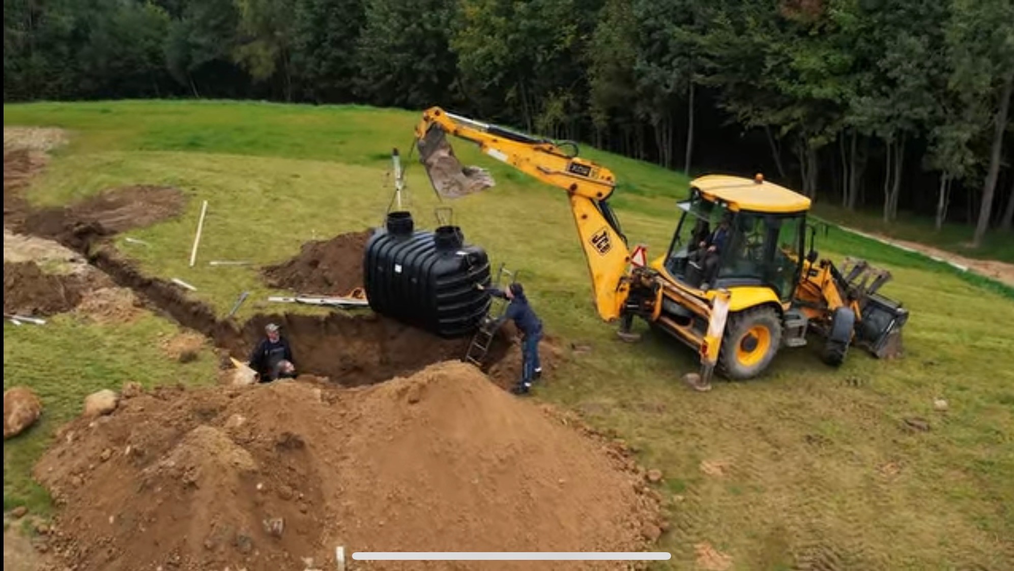 Instalacja przydomowej oczyszczalni ścieków: Żółta koparka JCB opuszcza czarny zbiornik na dół wykopu, gdzie pracownik czeka na drabinie, drugi pracownik w wykopie, tło: zielone drzewa i trawa.