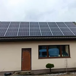 Nieduży, beżowy dom z brązowymi detalami i panelami fotowoltaicznymi na dachu, widoczne z zewnątrz przez okno niebieskie pojemniki.