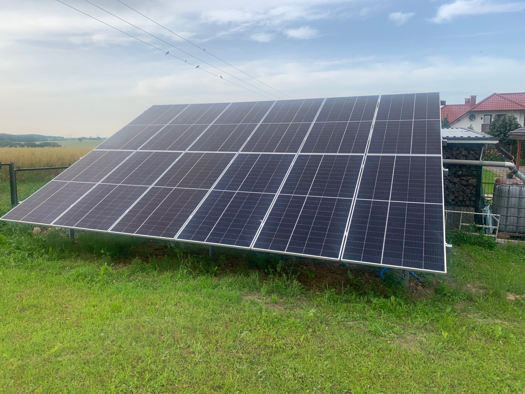 Instalacja paneli słonecznych na trawiastym terenie przydomowym; widoczne panele w metalowej ramie, w tle dom z czerwoną dachówką i pole uprawne.