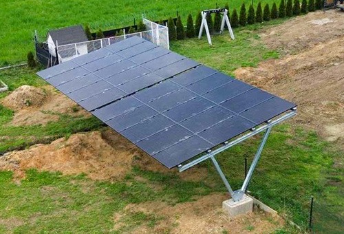 Instalacja fotowoltaiczna na gruncie, typu Carport V, z panelami Jinko, widoczna na tle zielonego trawnika i ogrodzenia z siatki, z małym budynkiem gospodarczym w tle.