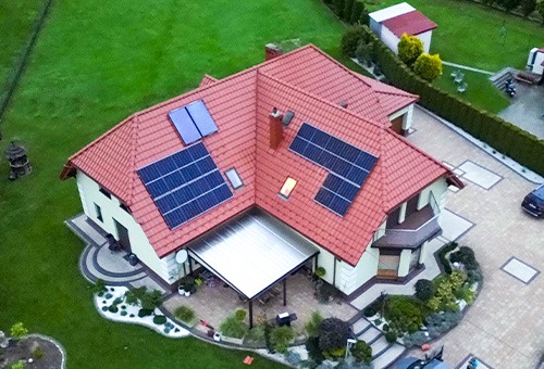 Widok z góry na dom jednorodzinny z czerwoną dachówką, na której zamontowano panele słoneczne; instalacja obejmuje kilka grup paneli, w tym dwie mniejsze grupy, a także widoczne świetliki dachowe.