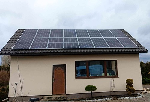 Nieduży, beżowy dom z brązowymi detalami i panelami fotowoltaicznymi na dachu, widoczne z zewnątrz przez okno niebieskie pojemniki.