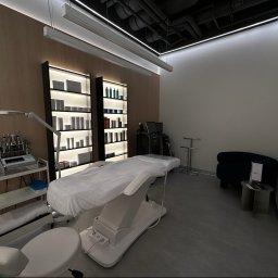 KOSMETOLOGIA ESTETYCZNA PIED-DE-POULE - Nowoczesny gabinet z regulowanym fotelem kosmetycznym przykrytym białym prześcieradłem, w tle regały z kosmetykami i sprzęt do zabiegów, oświetlenie sufitowe.