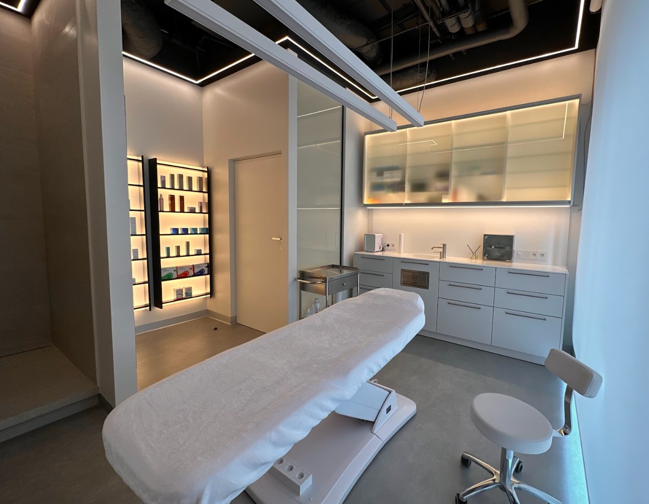 Jasne, minimalistyczne wnętrze gabinetu z leżanką kosmetyczną, szafkami z produktami i krzesłem na kółkach, oświetlone ledowymi lampami sufitowymi.