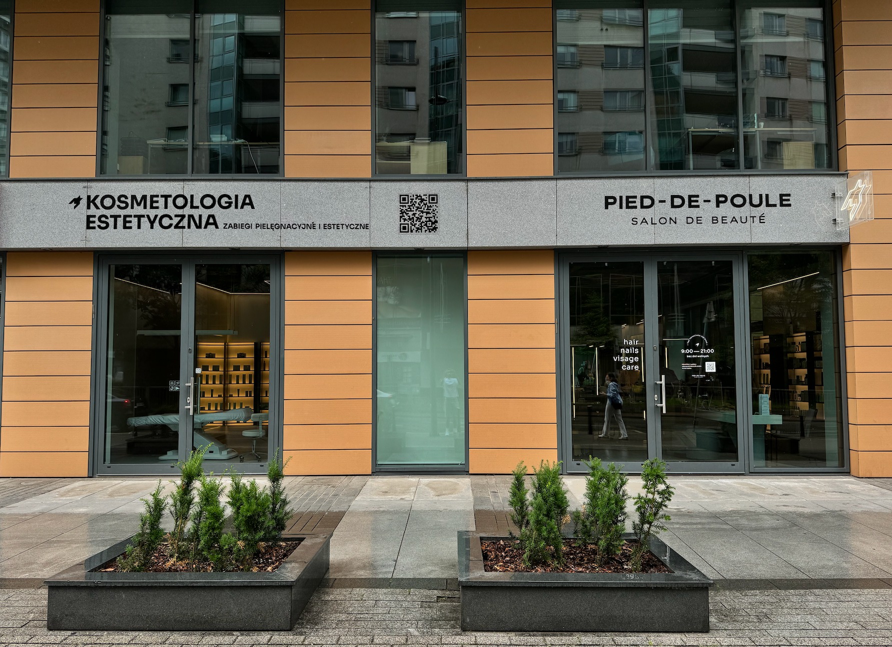 Fasada budynku z dwoma lokalami: 'Kosmetologia Estetyczna' z lewej i 'Pied-de-Poule Salon de Beauté' z prawej, przed wejściami donice z iglakami.