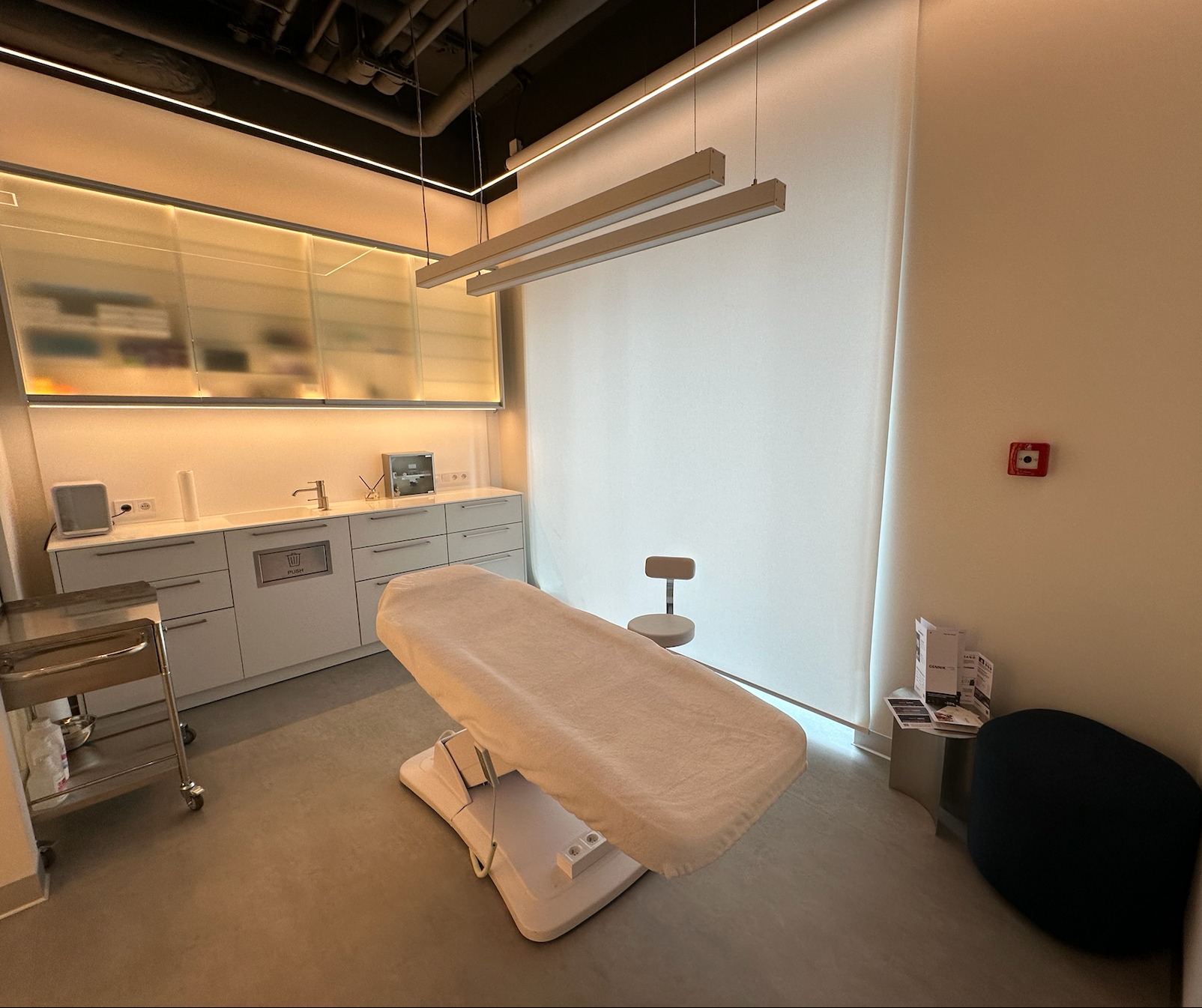 Jasne wnętrze gabinetu z regulowanym łóżkiem kosmetycznym przykrytym beżowym pokrowcem, minimalistycznym krzesłem, szafkami z wyposażeniem, oświetleniem LED i systemem wentylacji na suficie.