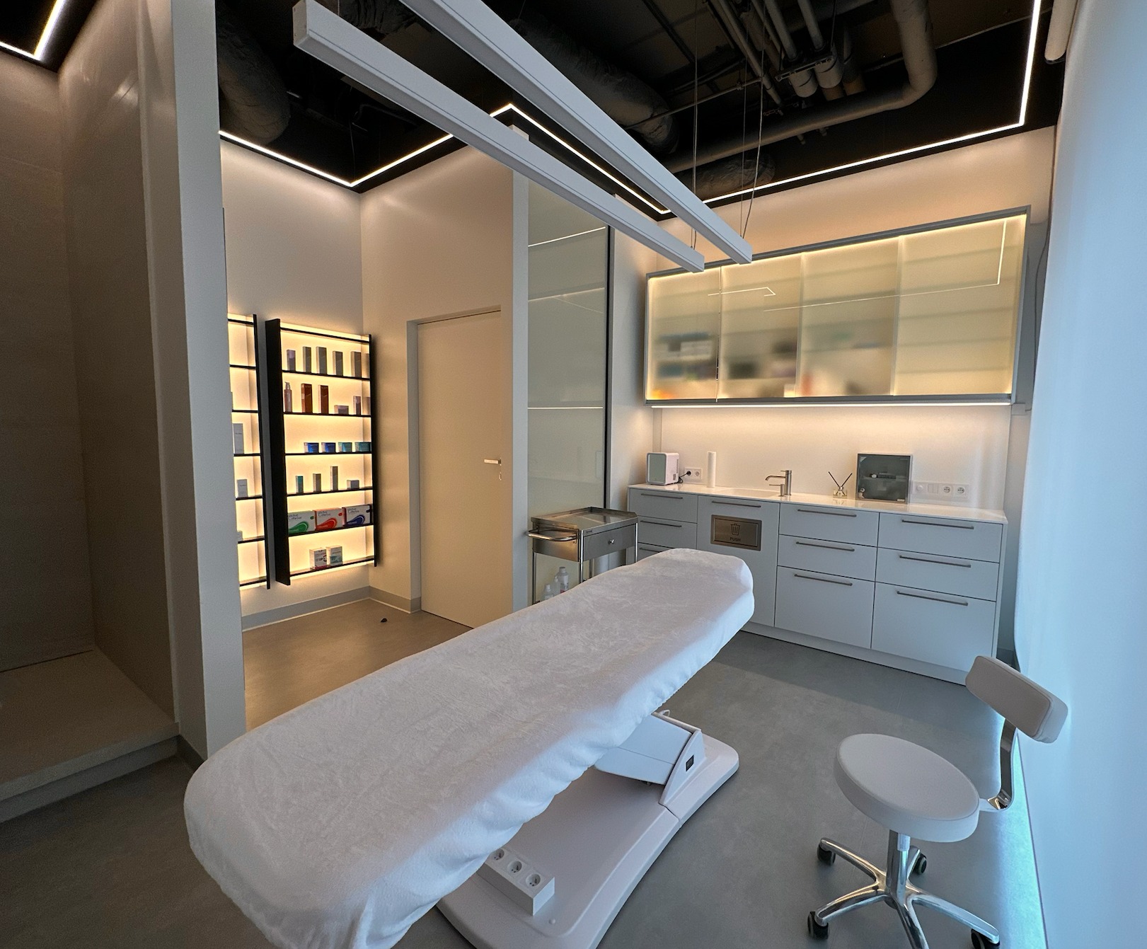 Jasne wnętrze gabinetu z leżanką kosmetyczną przykrytą białym prześcieradłem, obok krzesło na kółkach, w tle szafki z kosmetykami i umywalka, oświetlenie LED.