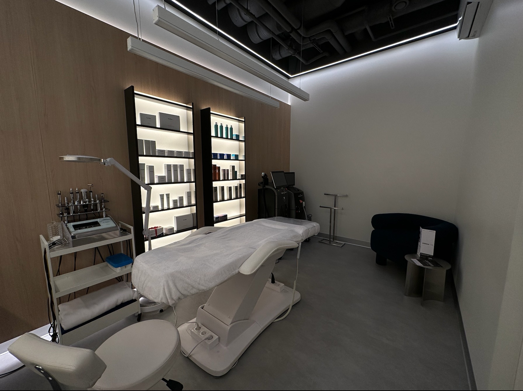 Nowoczesny gabinet z regulowanym fotelem kosmetycznym przykrytym białym prześcieradłem, w tle regały z kosmetykami i sprzęt do zabiegów, oświetlenie sufitowe.