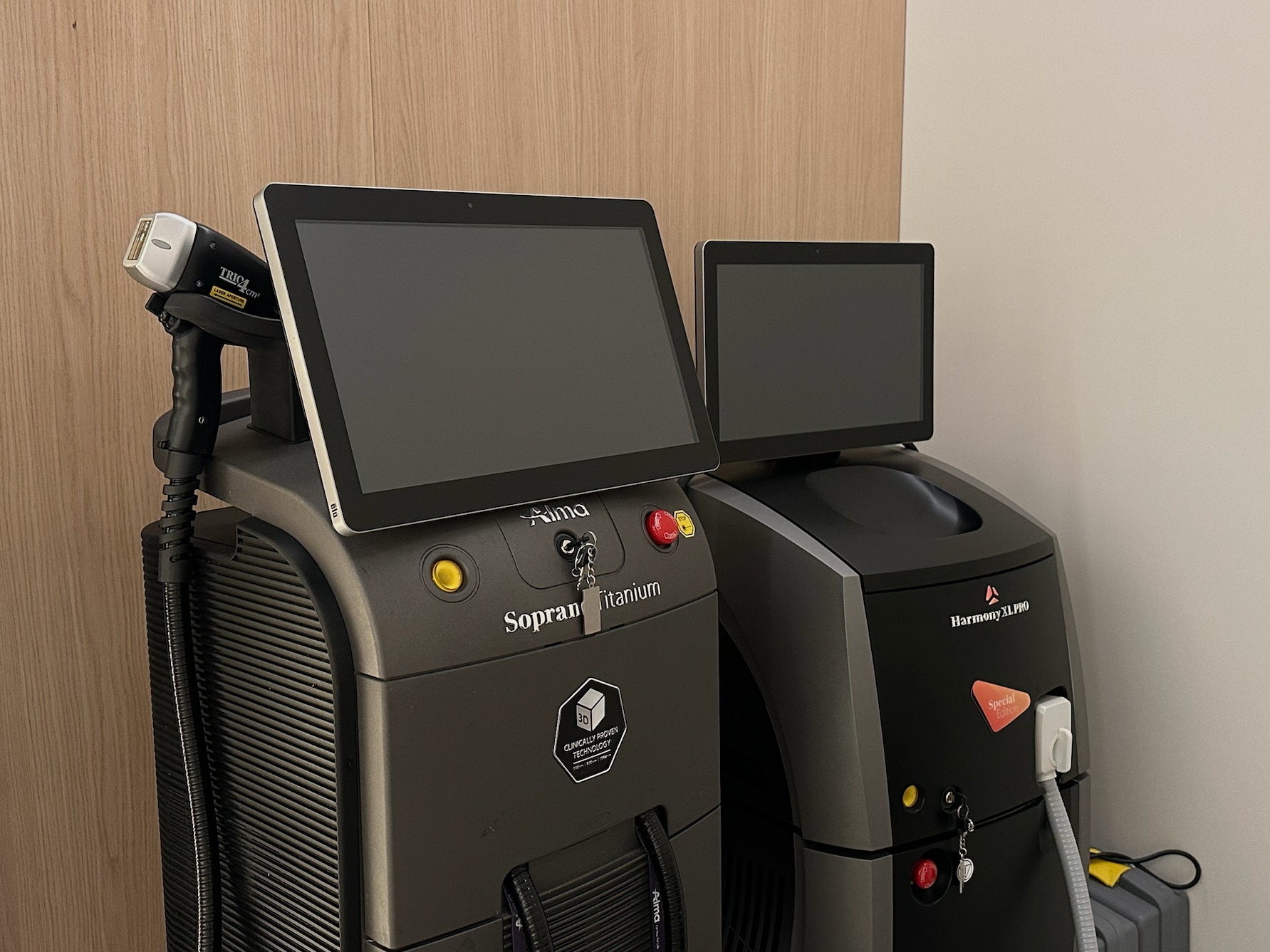 Dwa urządzenia do zabiegów laserowych, model Soprano Titanium i Harmony XL PRO, ustawione obok siebie w gabinecie, z widocznymi panelami dotykowymi i głowicą lasera.