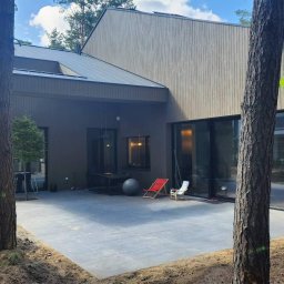 BrytanBudownictwo - Nowoczesny dom z elewacją z drewnianych paneli, widok na taras z szarych płyt, drzewa na pierwszym planie.