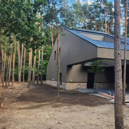 BrytanBudownictwo - Nowoczesny dom jednorodzinny z ciemną elewacją i oryginalnym dachem, otoczony lasem sosnowym, widok z poziomu gruntu na część frontową z podjazdem i fragmentem terenu przygotowanego pod trawnik.