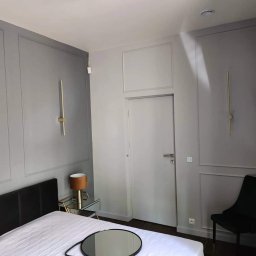 BrytanBudownictwo - Elegancka sypialnia w odcieniach szarości z dekoracyjnymi listwami ściennymi, złotymi lampami ściennymi, czarnym zagłówkiem łóżka i stolikiem nocnym z lustrzanym blatem.
