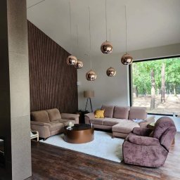 BrytanBudownictwo - Nowoczesny salon z drewnianą ścianą, oświetleniem kulistym w kolorze miedzi i dużym oknem z widokiem na las.