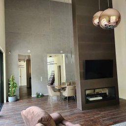 BrytanBudownictwo - Wysokie wnętrze domu w Słopnicach z szarą ścianą wykończoną płytami, widoczne drzwi przesuwne, drewniana podłoga, nowoczesny kominek z telewizorem i wiszące lampy kuliste.