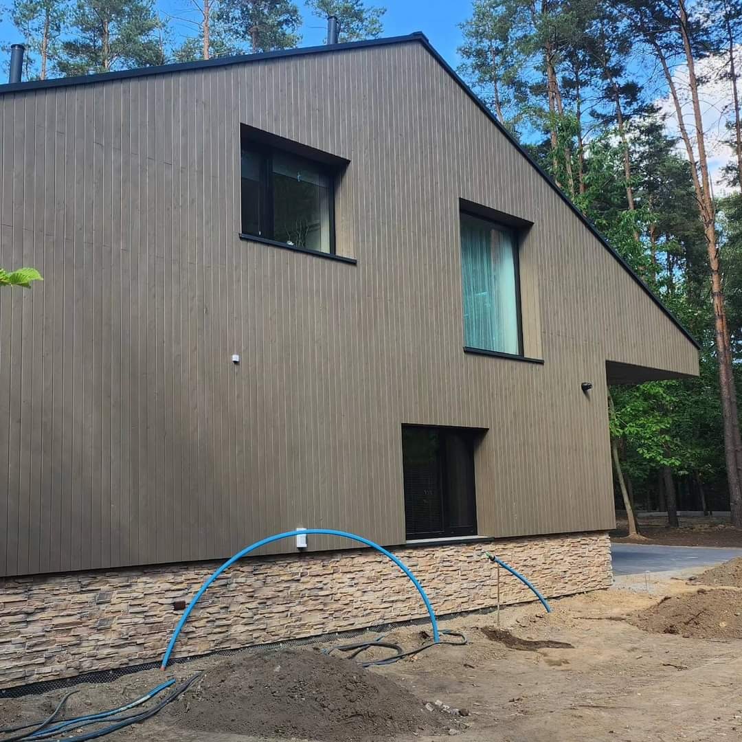 Nowoczesny dom murowany z drewnianą elewacją w odcieniach brązu i kamiennym cokołem, widoczne niebieskie rury na zewnątrz, teren wokół domu w trakcie prac ziemnych.