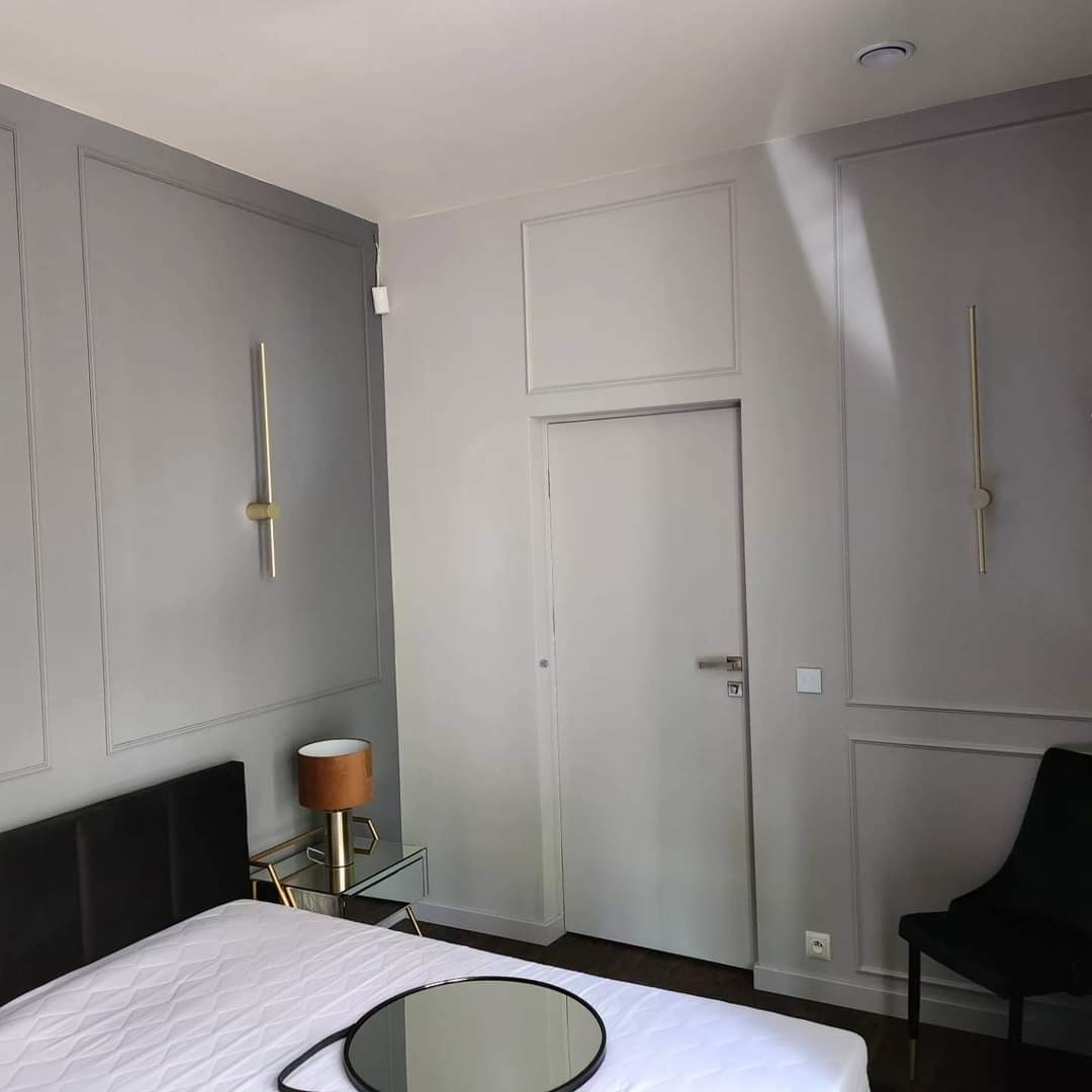 Elegancka sypialnia w odcieniach szarości z dekoracyjnymi listwami ściennymi, złotymi lampami ściennymi, czarnym zagłówkiem łóżka i stolikiem nocnym z lustrzanym blatem.
