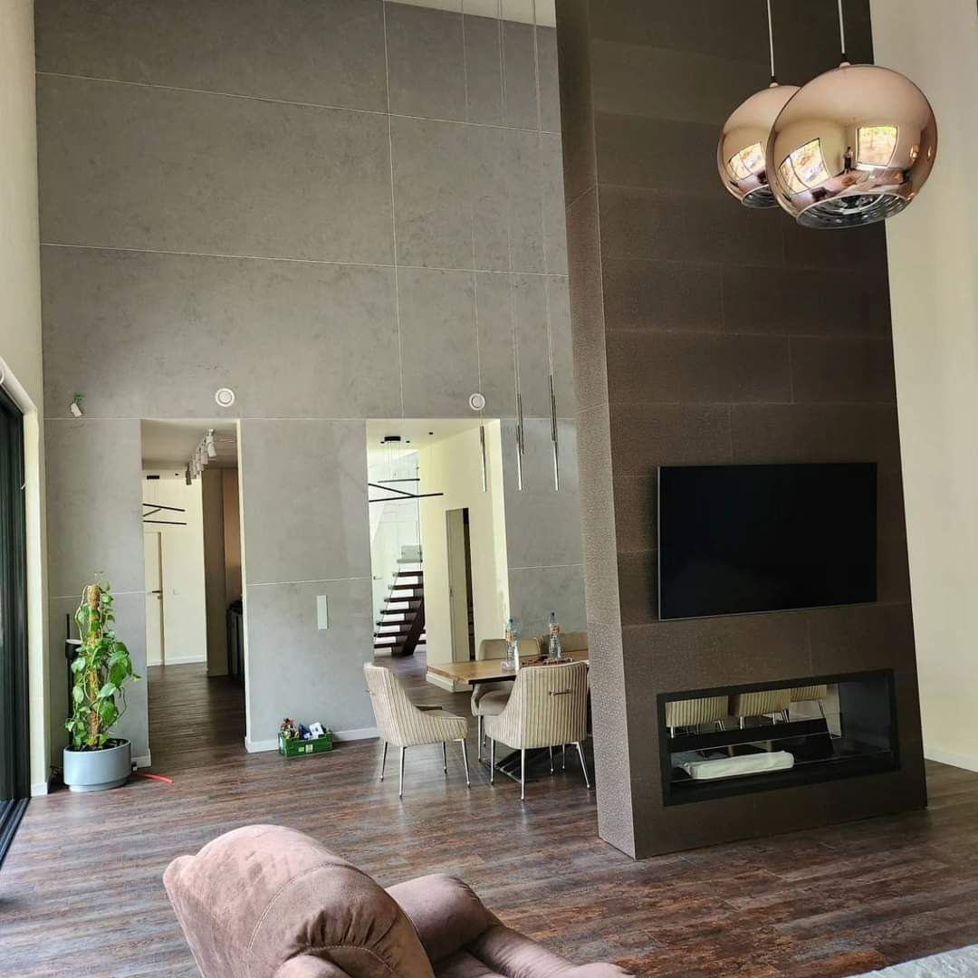 Wysokie wnętrze domu w Słopnicach z szarą ścianą wykończoną płytami, widoczne drzwi przesuwne, drewniana podłoga, nowoczesny kominek z telewizorem i wiszące lampy kuliste.