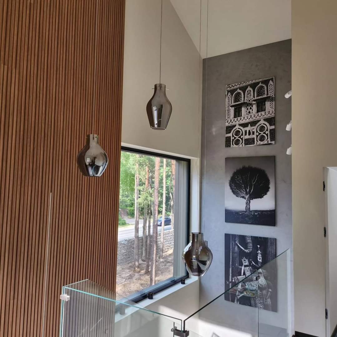 Nowoczesna klatka schodowa z drewnianą ścianą, szklaną balustradą, wiszącymi lampami w kształcie wazonów i grafikami na szarej ścianie w tle.