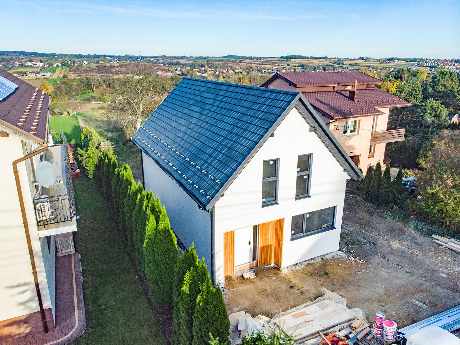 Nowoczesny dom szkieletowy z ciemnym dachem, białymi ścianami i drewnianymi drzwiami wejściowymi, widok z góry na tle zielonego krajobrazu i sąsiednich domów.