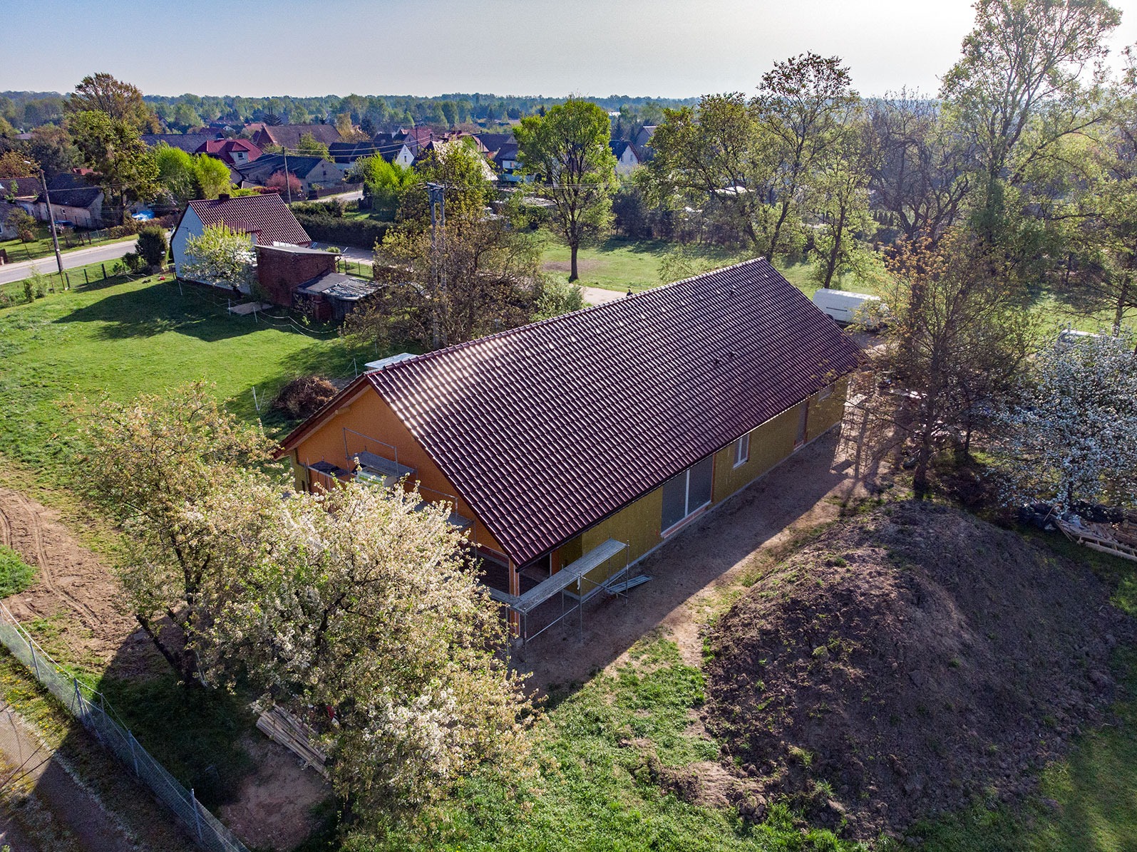 Wznoszony parterowy budynek z ciemnoczerwoną dachówką, widok z lotu ptaka, rusztowania przy ścianie bocznej, drzewa owocowe w kwitnieniu.