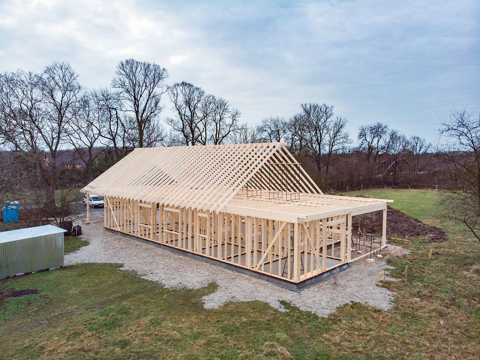 Konstrukcja drewniana domu w trakcie budowy, widok z lotu ptaka, z widoczną więźbą dachową i ścianami szkieletowymi, na zewnątrz krajobraz z drzewami bez liści i fragmentem nieba.