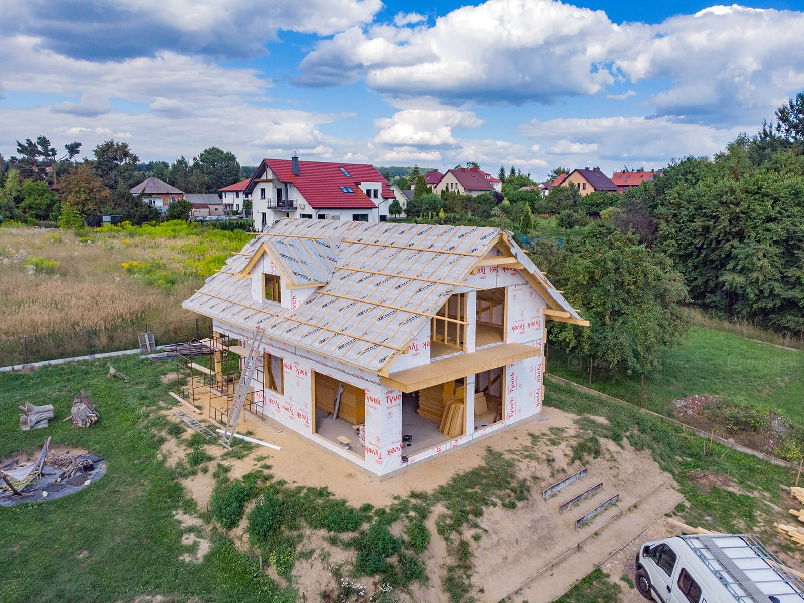 Budowa domu w Leśnej w trakcie realizacji. Widok z góry na dwukondygnacyjny dom w stanie surowym otwartym, zabezpieczony folią izolacyjną Tyvek, z widocznym drewnianym szkieletem dachu i ścian,...