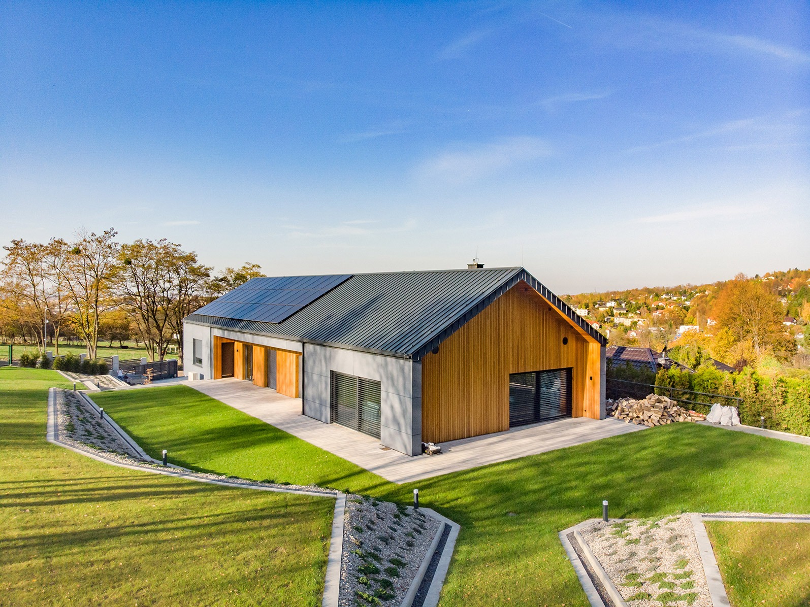 Nowoczesny dom z panelami słonecznymi na dachu, widok z góry na zadbany ogród z elementami małej architektury i otaczający krajobraz.