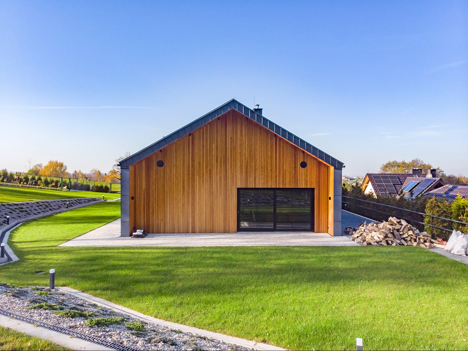 Nowoczesny dom z drewnianą elewacją i grafitowym dachem na tle zielonego trawnika i błękitnego nieba, z widocznym stosem drewna obok.