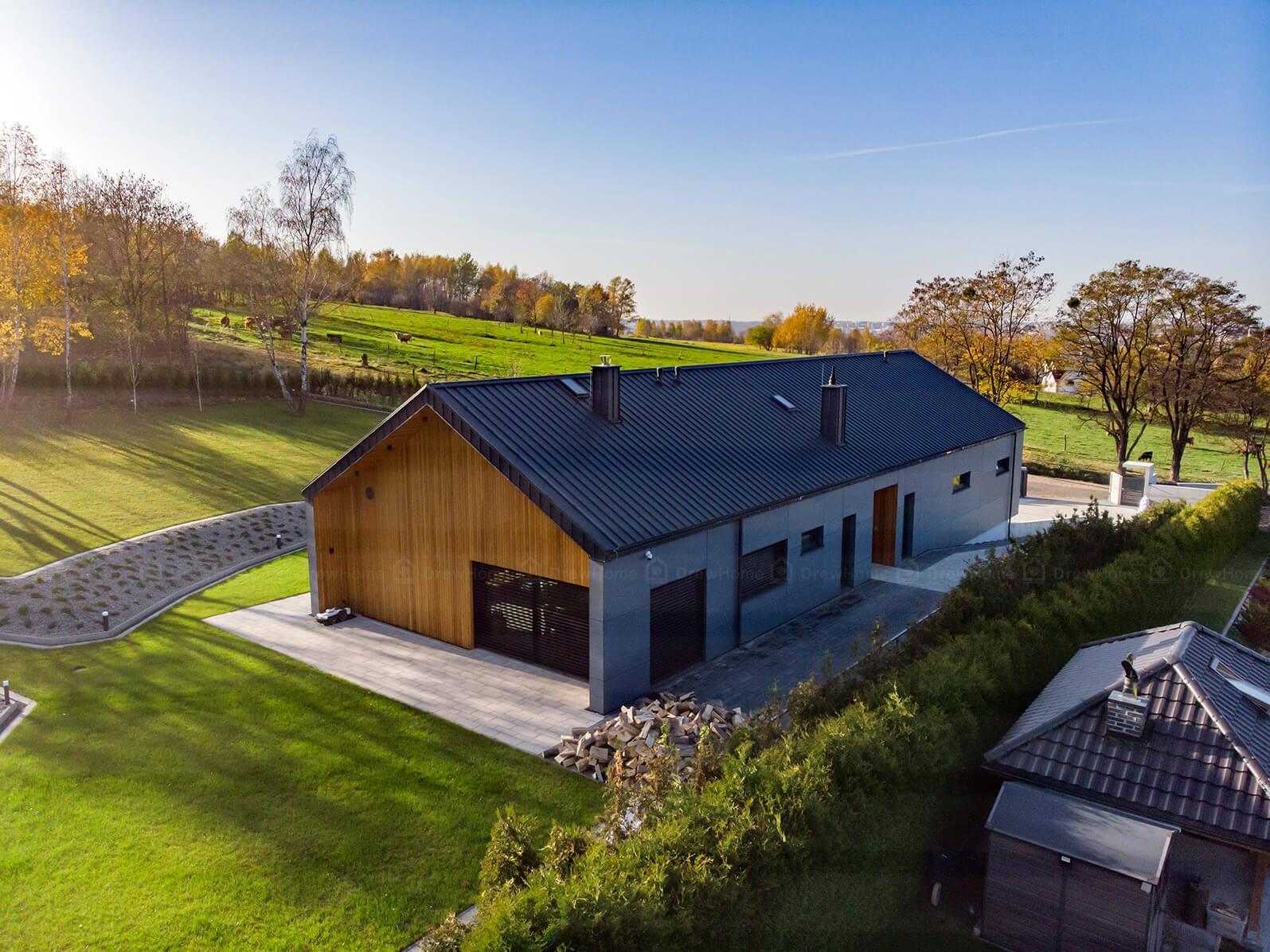 Nowoczesny dom z drewnianą elewacją i ciemnym dachem na tle zielonego krajobrazu z pasącymi się zwierzętami, widok z lotu ptaka.