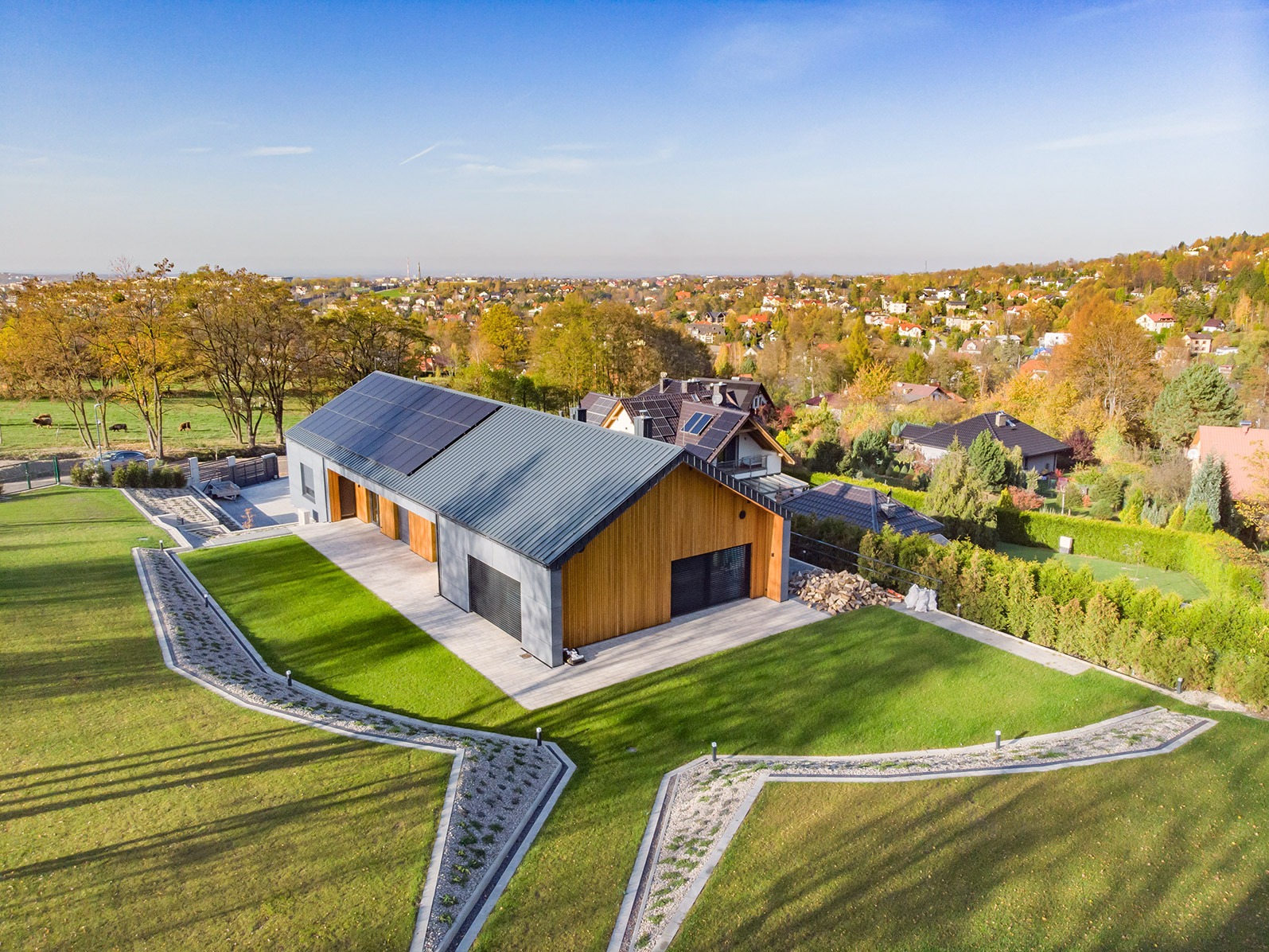Nowoczesny dom szkieletowy z panelami słonecznymi, widok z lotu ptaka na zadbany ogród i okoliczną zabudowę.