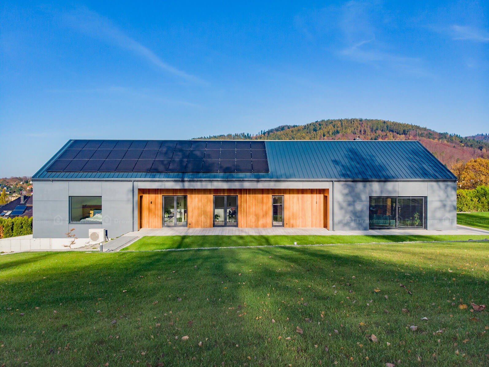 Nowoczesny dom z panelami słonecznymi na dachu, wykończony drewnem i szarym sidingiem, otoczony zielonym trawnikiem i jesiennym krajobrazem.