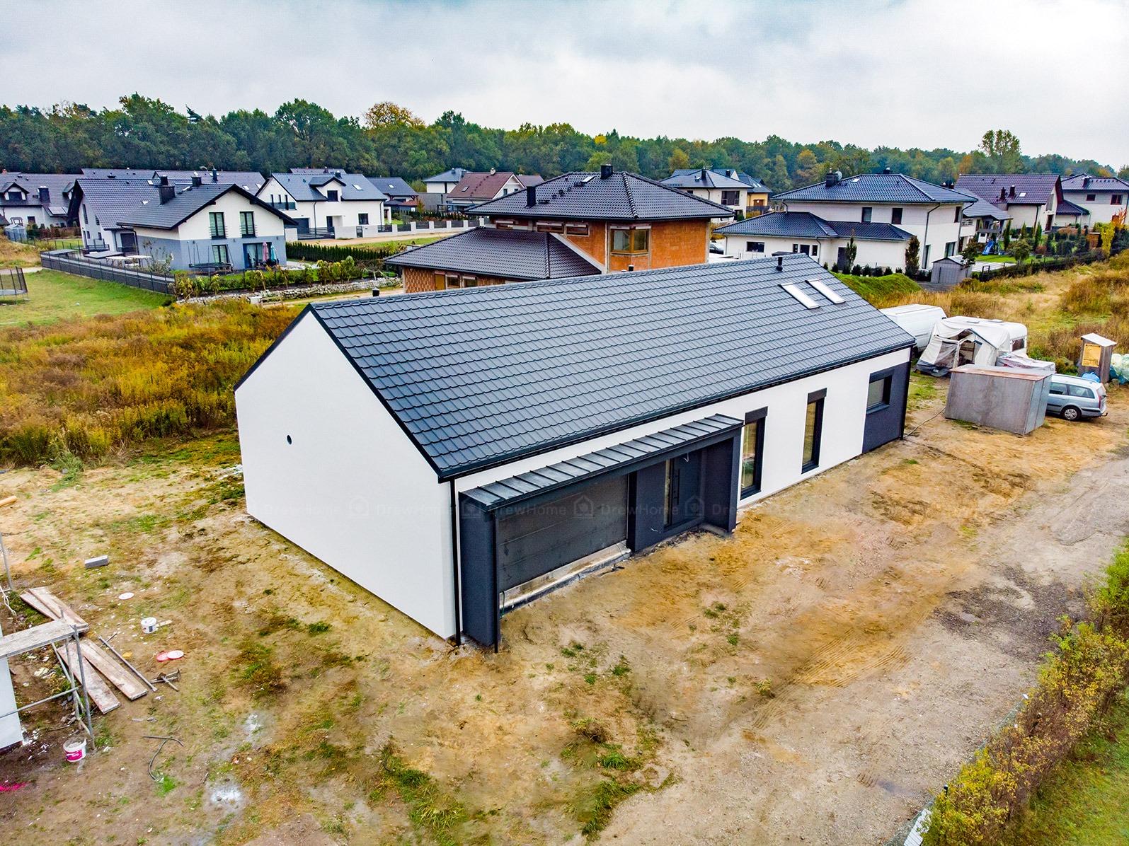 Nowoczesny, parterowy dom z ciemnoszarą dachówką i białymi ścianami, widoczny z lotu ptaka na tle innych domów i drzew, z elementami placu budowy w otoczeniu.