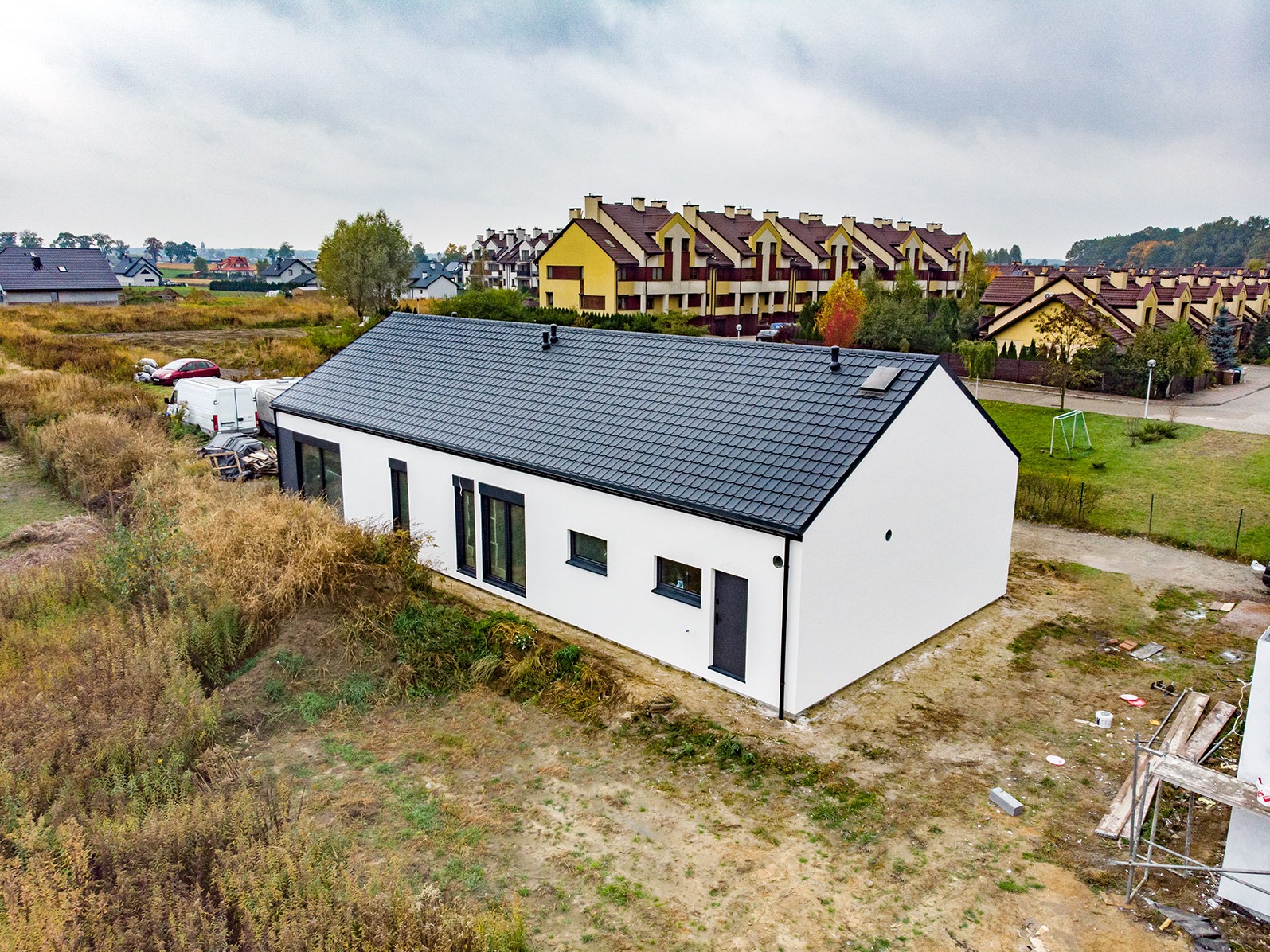 Nowoczesny, biały dom jednorodzinny z czarnym dachem i stolarką okienną, widok z góry na niezagospodarowaną działkę, w tle zabudowa szeregowa.