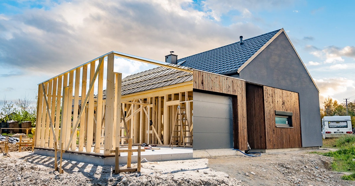 Budowa domu: widok na konstrukcję szkieletową garażu z przylegającą do niego częścią mieszkalną z szarą elewacją i drewnianymi panelami, drabina oparta o ścianę, w tle przyczepa kempingowa.