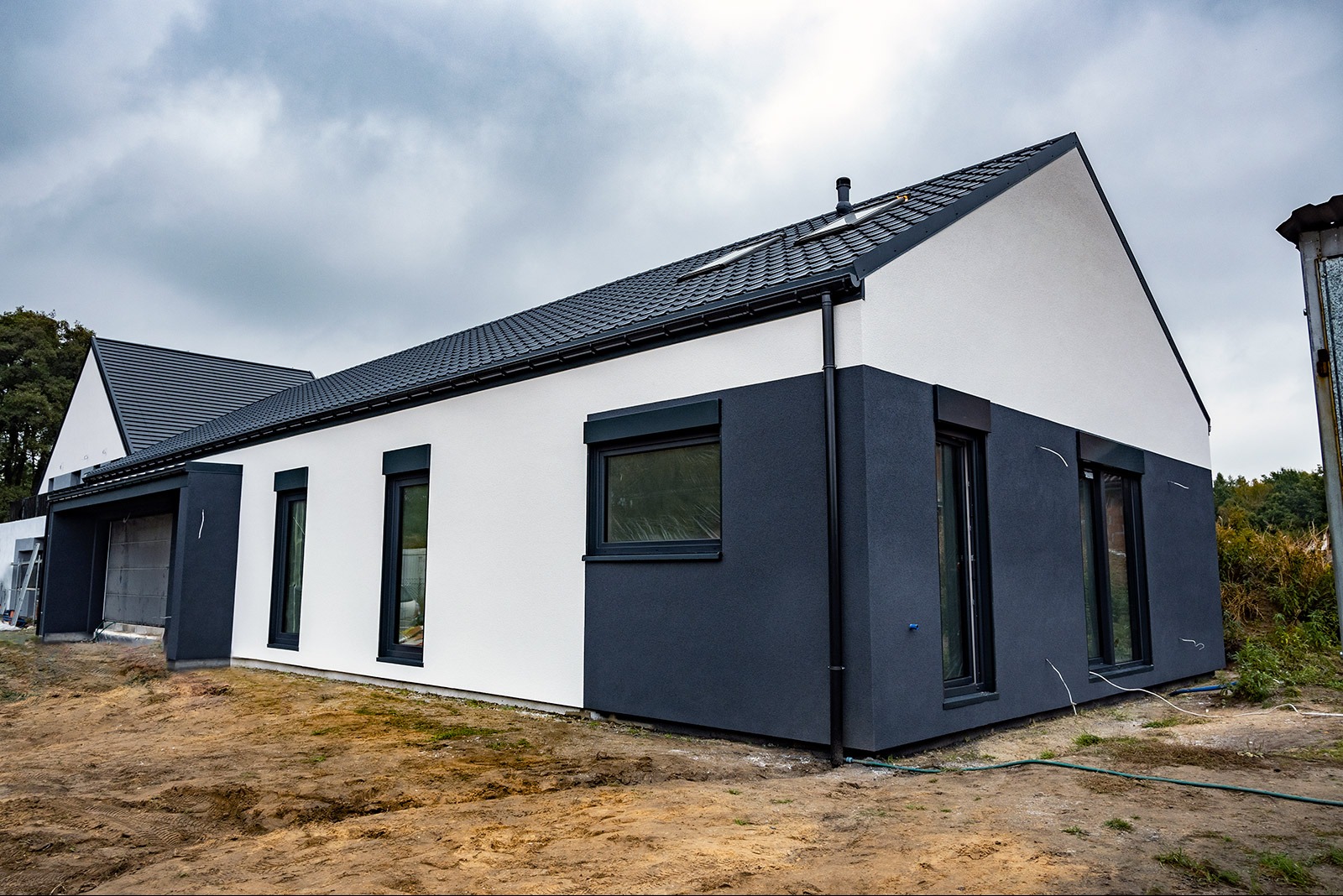 Nowoczesny dom z elementami szkieletowymi w trakcie wykończenia, z dwukolorową elewacją (biało-szara), ciemną dachówką i czarnymi ramami okiennymi; widoczne okno dachowe i niezagospodarowany teren...