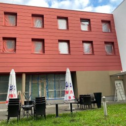JC- Remonty i wykończenia - Czerwona elewacja budynku w trakcie malowania. Okna zabezpieczone folią, widoczny trawnik z parasolami i meblami ogrodowymi.