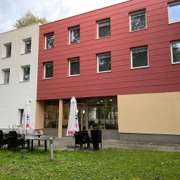 JC- Remonty i wykończenia - Odnowiona elewacja budynku z czerwonymi i białymi panelami, widoczne okna z białymi ramami i fragment zewnętrznego ogródka. Świeżo pomalowana fasada.