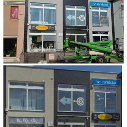 JC- Remonty i wykończenia - Odnowiona fasada budynku z szarą i czarną elewacją. Widoczne okna z reklamami firm, podnośnik koszowy i detale architektoniczne. Dzień, światło słoneczne.