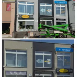 Odnowiona fasada budynku z szarą i czarną elewacją. Widoczne okna z reklamami firm, podnośnik koszowy i detale architektoniczne. Dzień, światło słoneczne.