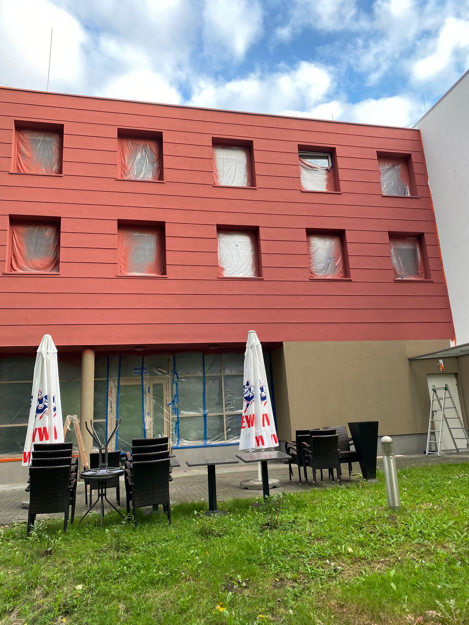 Czerwona elewacja budynku w trakcie malowania. Okna zabezpieczone folią, widoczny trawnik z parasolami i meblami ogrodowymi.