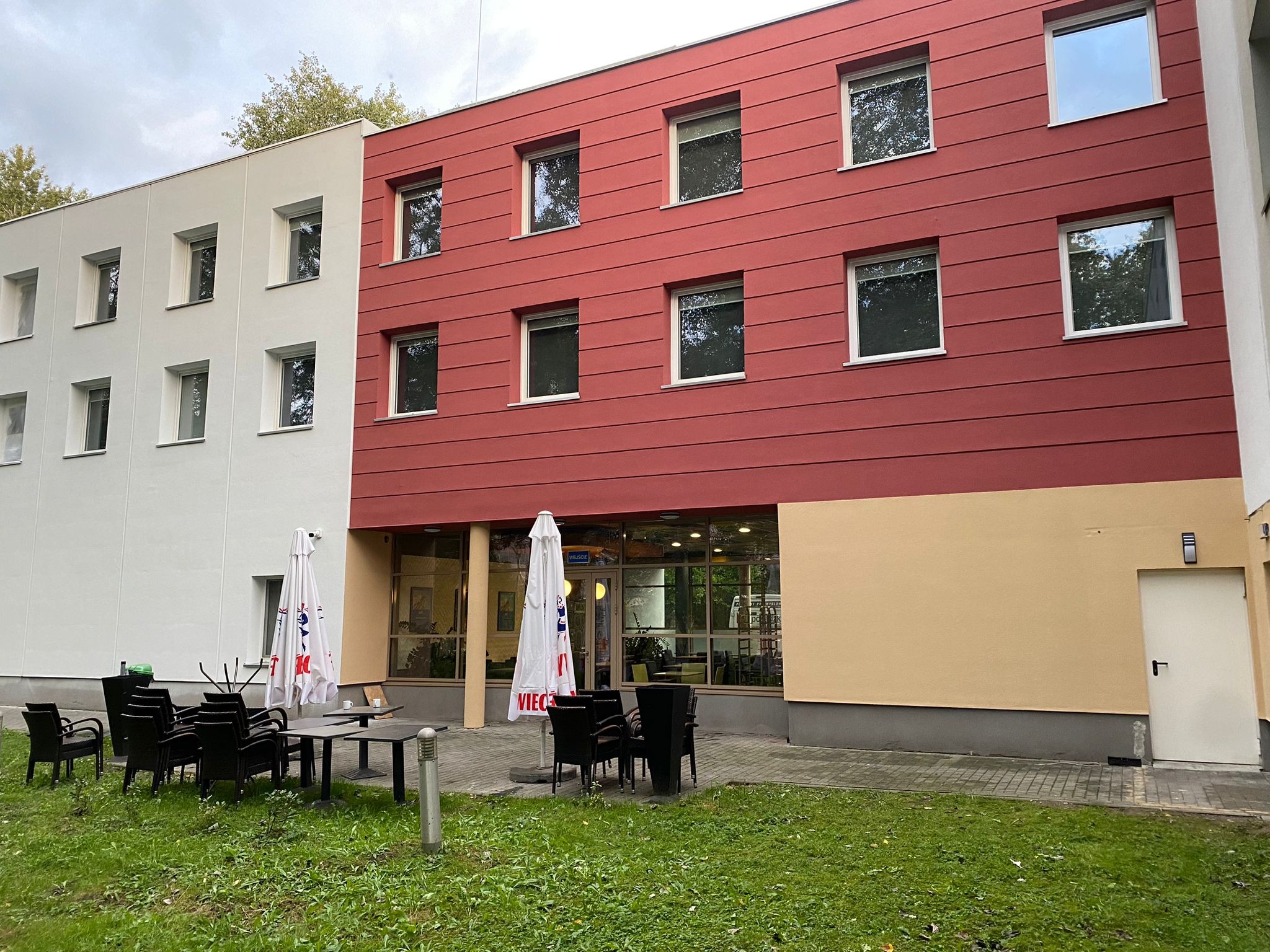 Odnowiona elewacja budynku z czerwonymi i białymi panelami, widoczne okna z białymi ramami i fragment zewnętrznego ogródka. Świeżo pomalowana fasada.