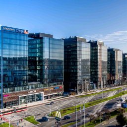 M-PROJECT SPÓŁKA Z OGRANICZONĄ ODPOWIEDZIALNOŚCIĄ - Panorama nowoczesnego kompleksu biurowego z przeszklonymi fasadami, widok z góry na ulicę z ruchem samochodowym i pieszym, w tle budowa kolejnego budynku z dźwigiem.