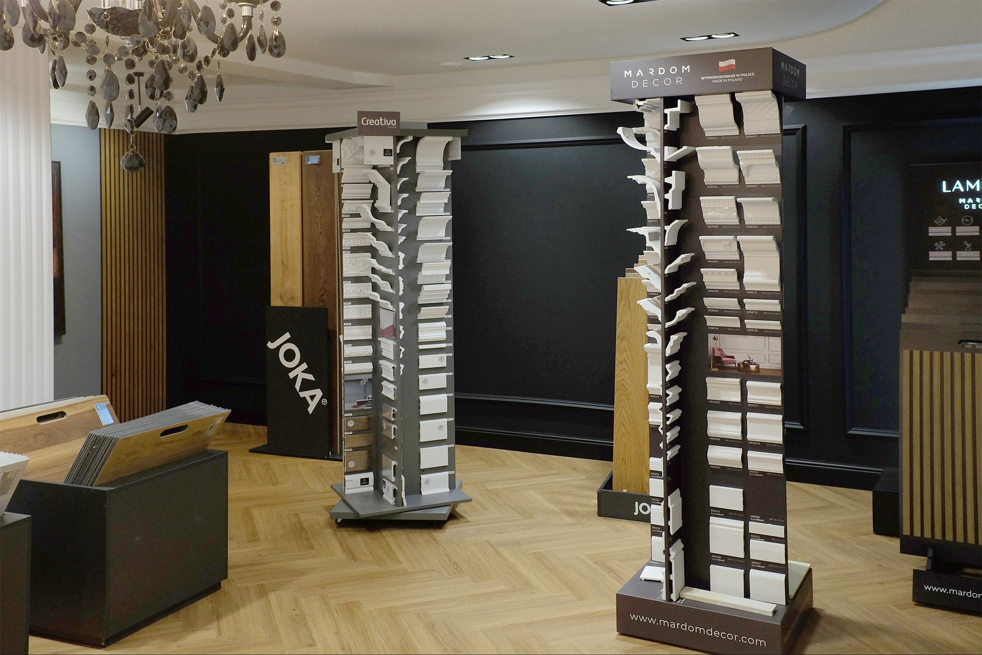 Ekspozycja sztukaterii wewnętrznej marek Mardom Decor i Creativa na stojakach w salonie, widoczne listwy przypodłogowe, sufitowe i ścienne różnych wzorów oraz panele podłogowe Joka.